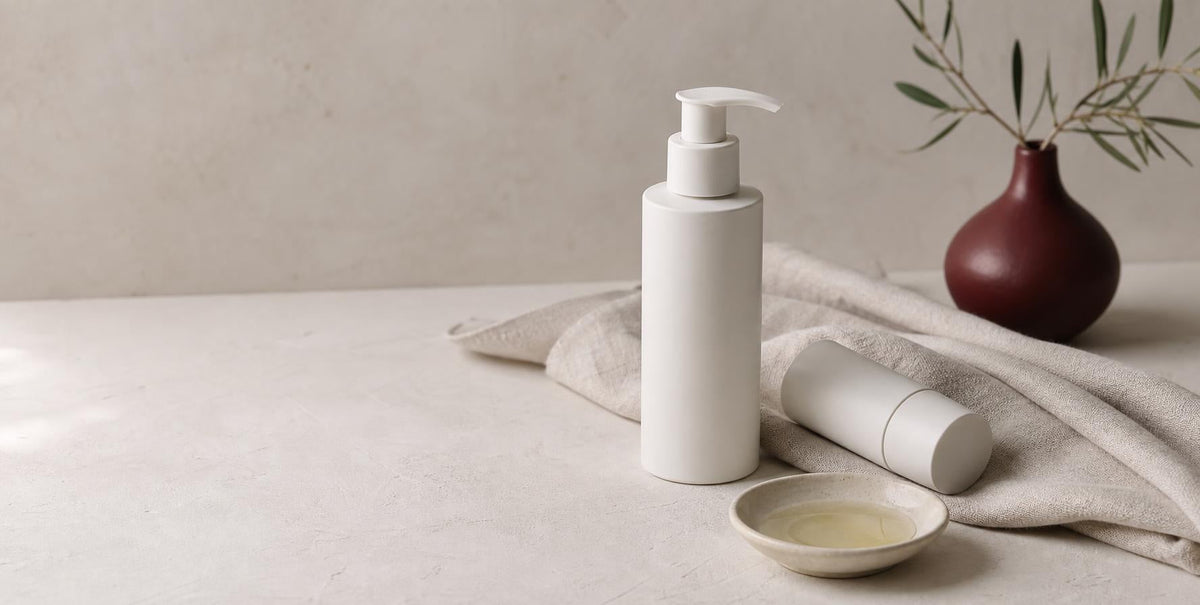 White pump bottle and tube beside a small dish of oil on linen, illustrating fragrance-free skincare formulation